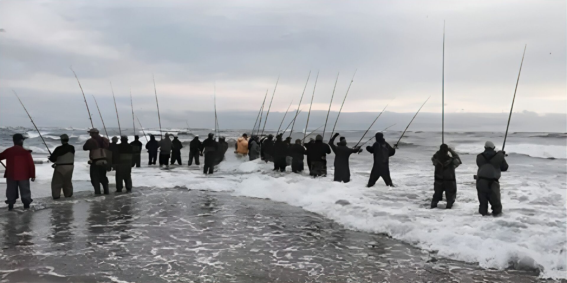 Group of people fishing together at the shoreline under a cloudy sky.