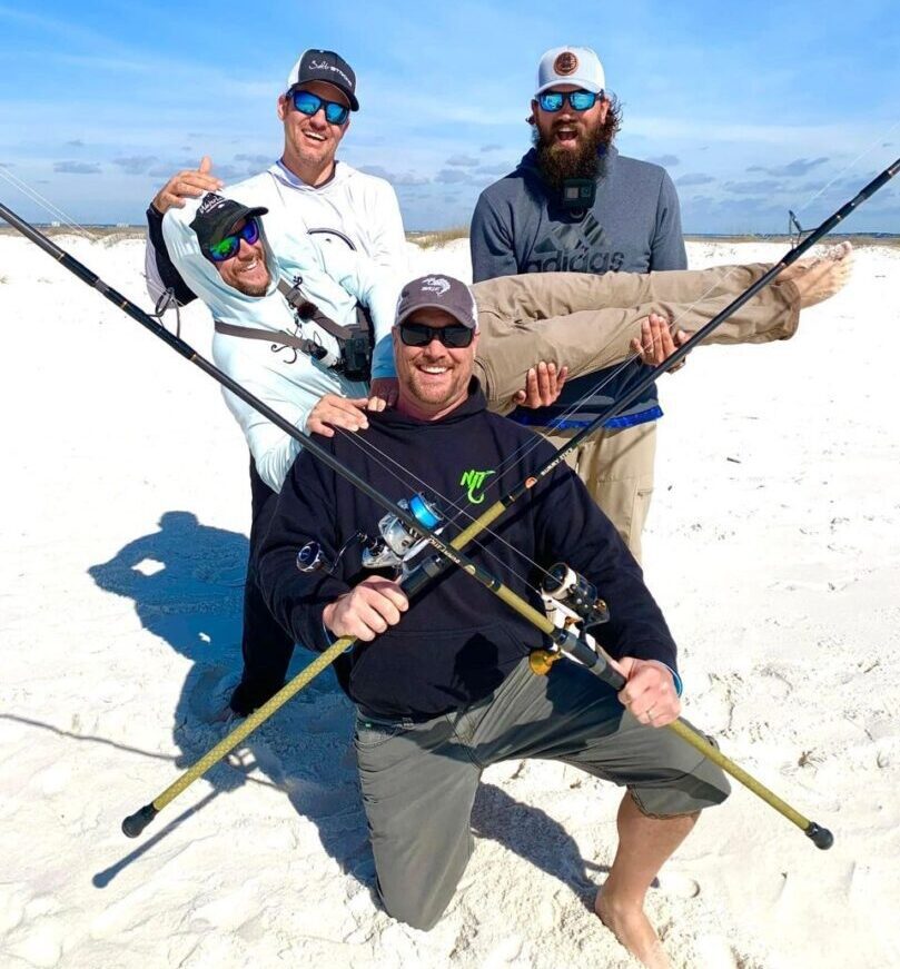 Four men posing with fishing rods on a sandy beach under clear skies.
